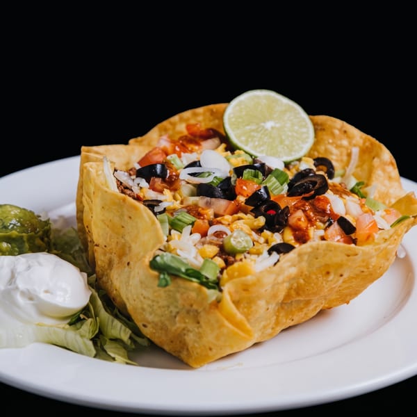 Taco salad in a crispy tortilla bowl with ground beef, vegetables, cheese, and a lime wedge.