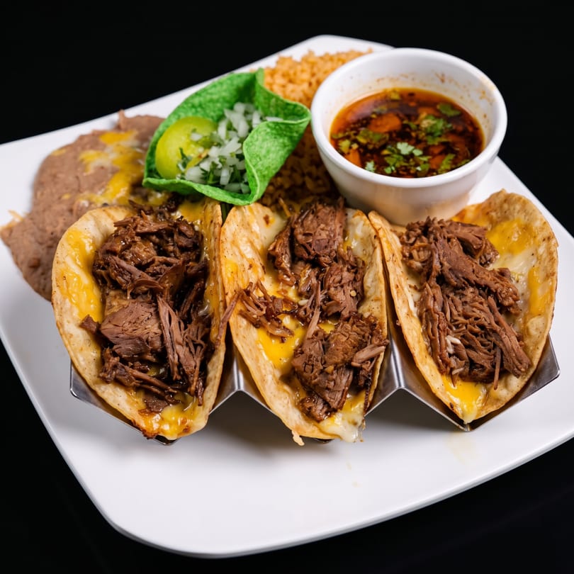 Three birria tacos with melted cheese and shredded beef, served with rice, refried beans, and consommé.